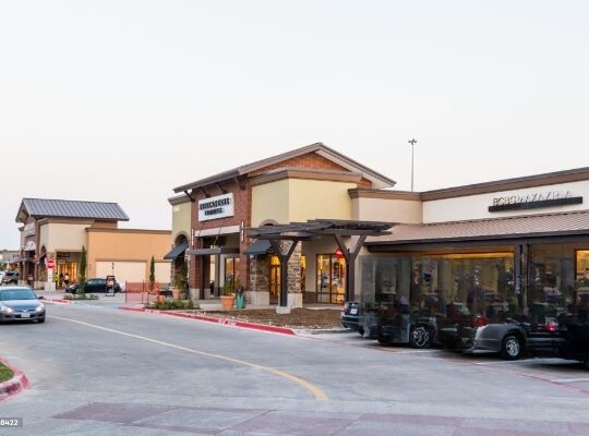 Distant view of Allen Premium Outlets in Dallas, Texas, showcasing the outdoor shopping complex surrounded by green spaces and featuring a variety of retail stores.