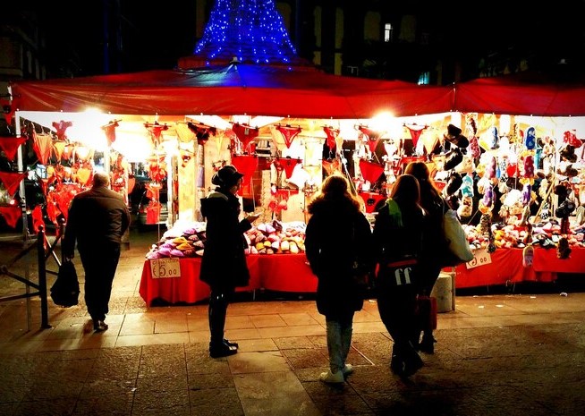 "Shoppers explore a festive holiday market at night, illuminated by vibrant red and warm lights with a decorated Christmas tree in the background." one of the activity from the Dallas winter bucket.