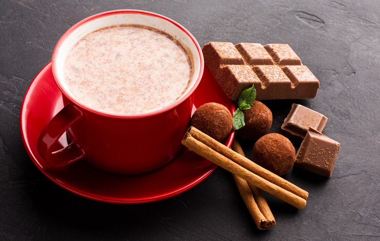 A red mug filled with a hot, creamy beverage surrounded by an assortment of chocolate confections and spices