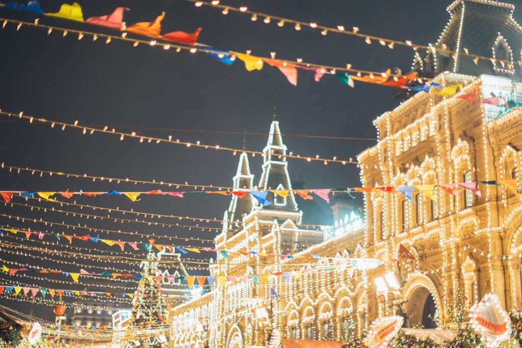 A historic, ornate building illuminated by strands of colorful lights, with rows of vibrant flags or banners suspended in the foreground, creating a festive, celebratory night scene